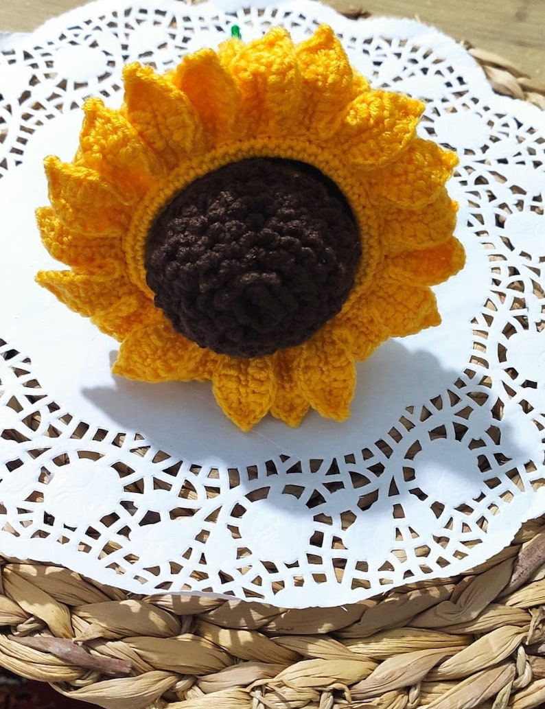 K&ouml;nnte beinhalten: Eine geh&auml;kelte Sonnenblume mit gelben Bl&uuml;tenbl&auml;ttern und einer braunen Mitte. Die Sonnenblume liegt auf einer wei&szlig;en Spitze.