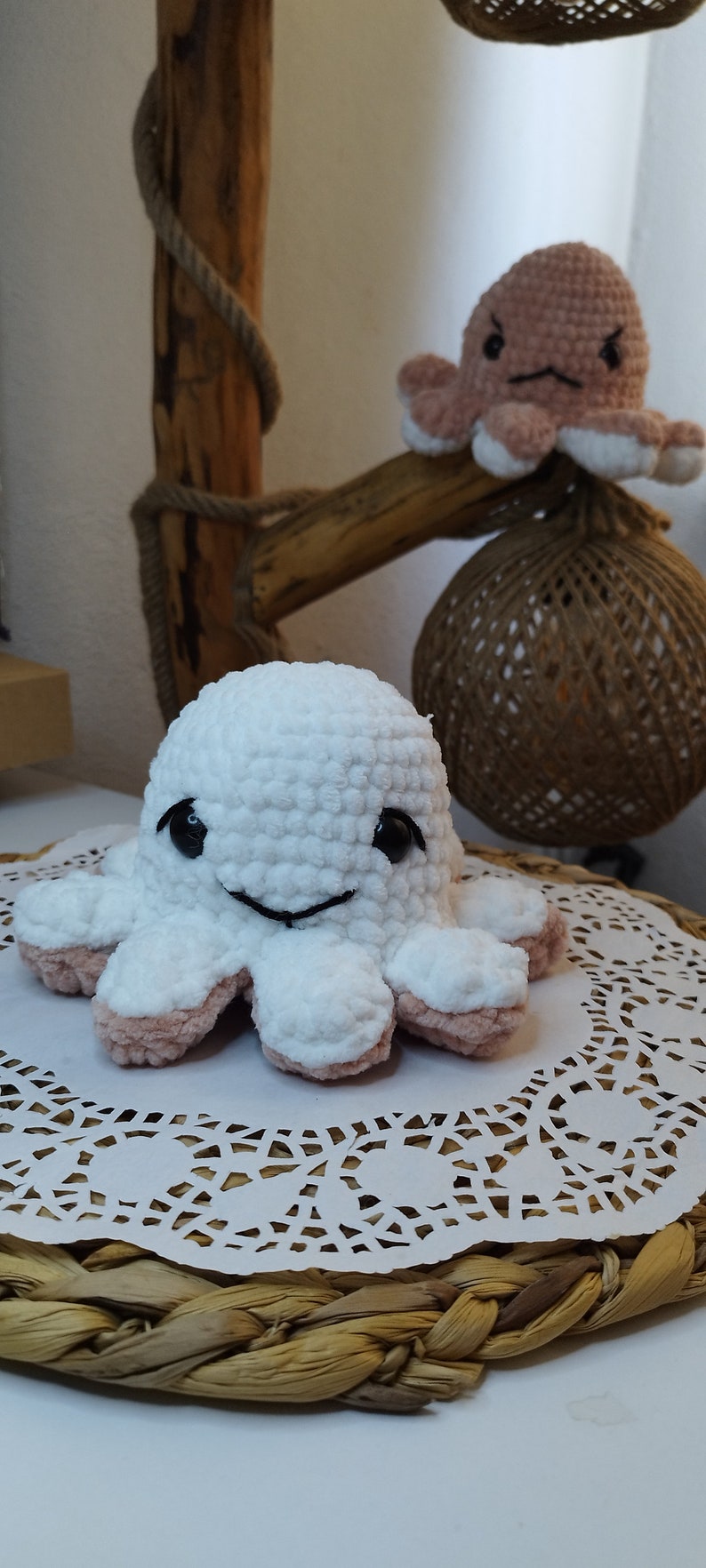 May include: A white crocheted octopus with pink accents and black eyes sits on a white doily on a woven basket.