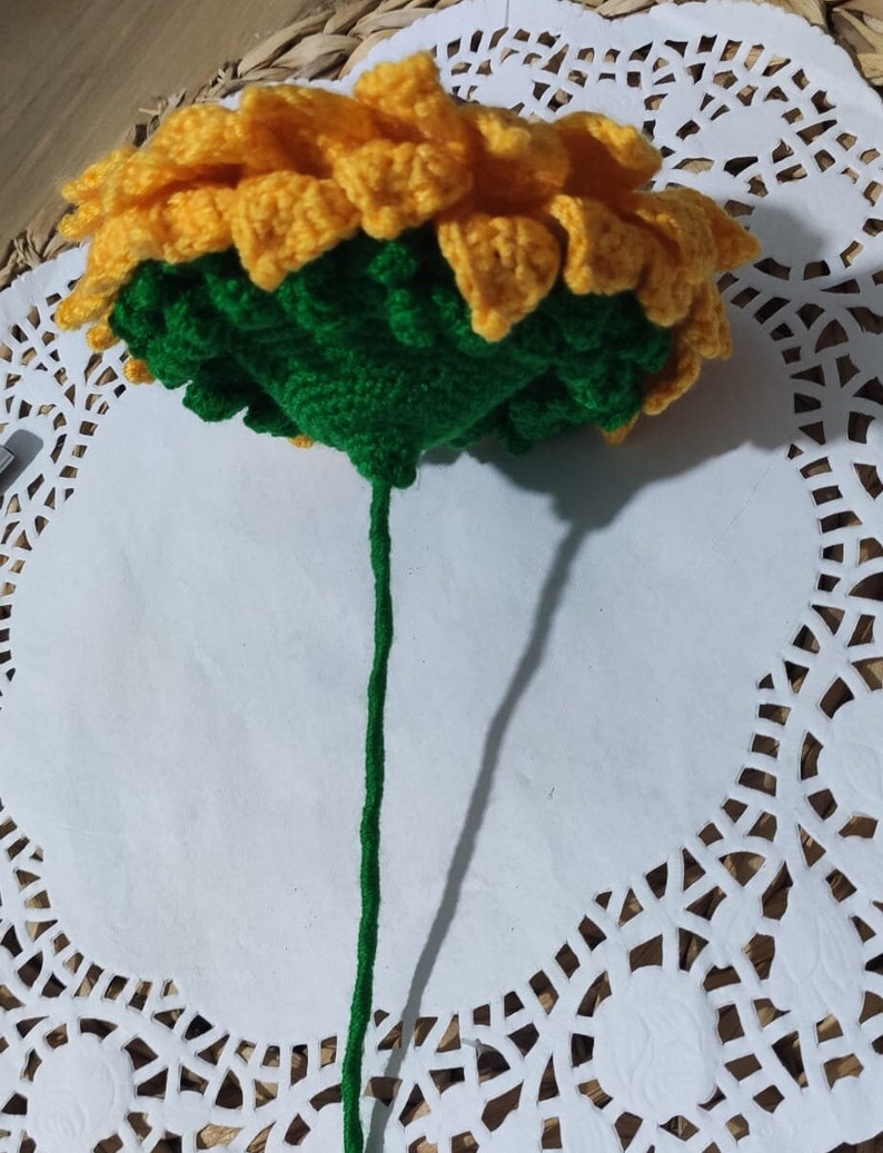 K&ouml;nnte beinhalten: Eine geh&auml;kelte Sonnenblume mit gelben Bl&uuml;tenbl&auml;ttern und einer gr&uuml;nen Mitte, die an einem gr&uuml;nen Stiel befestigt ist.
