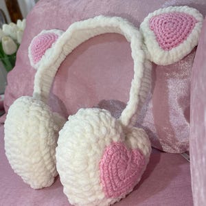 May include: White crocheted ear warmers with pink heart accents. The ear warmers are shaped like hearts and have a headband.