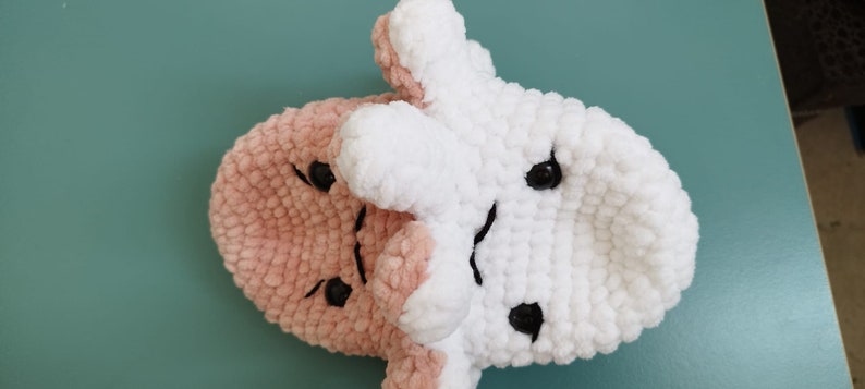 May include: A crocheted plush toy with a pink and white face. The toy has two black eyes and a black mouth.