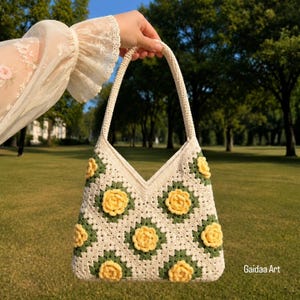 Può includere: Una borsa all'uncinetto con base color crema, un motivo ripetuto di quadrati verde scuro e decorazioni floreali gialle tridimensionali. La borsa presenta un manico singolo integrato e un'apertura superiore a V. Il testo Gaidaa Art è visibile.