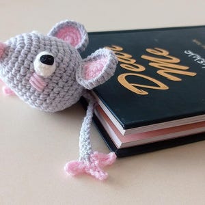 May include: A gray crocheted mouse bookmark with pink ears and nose, resting on a book. The mouse has a white eye and black pupil. The book has gold lettering on a dark cover.