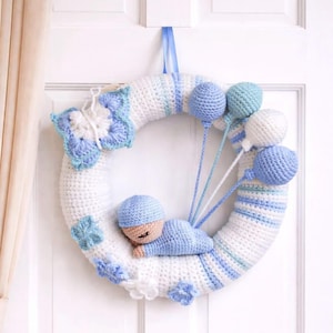 May include: A handmade crochet wreath in white and blue, decorated with a baby, balloons, a butterfly, and flowers. The wreath is suspended by a blue ribbon. The baby is wearing a blue hat and is lying on a blue cloud.