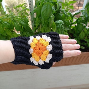 May include: Black crochet fingerless glove with a colourful floral design. The flower has an orange centre, surrounded by yellow and white petals. The glove is shown on a hand, with green plants in the background.