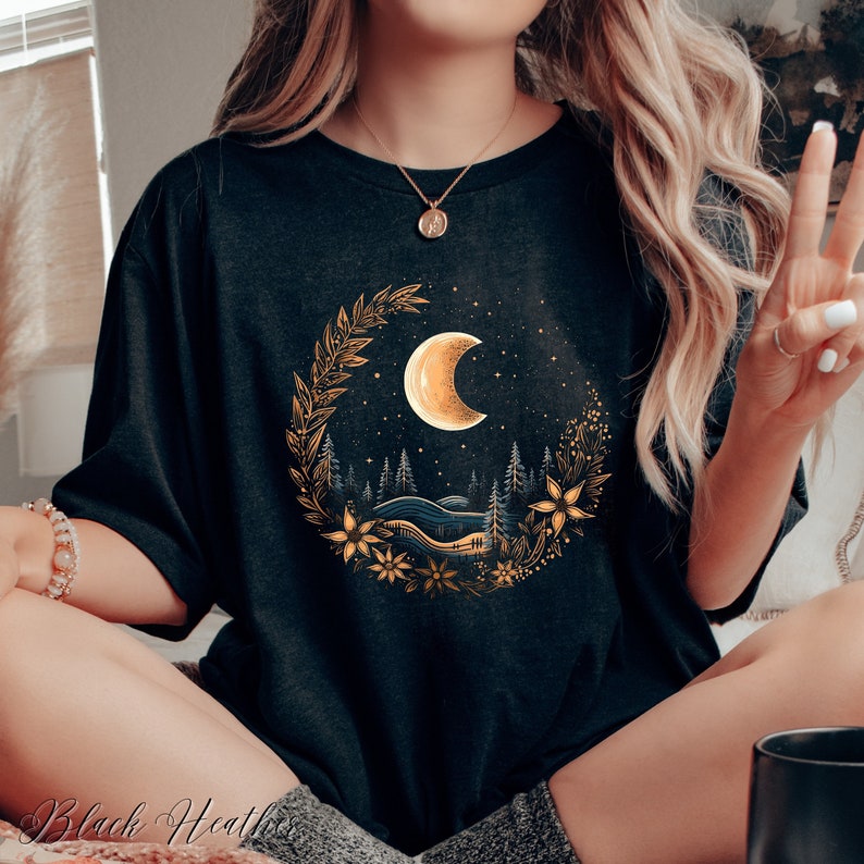 May include: A black t-shirt with a gold crescent moon and forest scene graphic. The moon is surrounded by a wreath of leaves and flowers.
