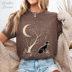 May include: Heather Brown t-shirt featuring a gold crescent moon, tree, and a black cat design. The graphic includes a starry night sky. The shirt is a neutral brown color.