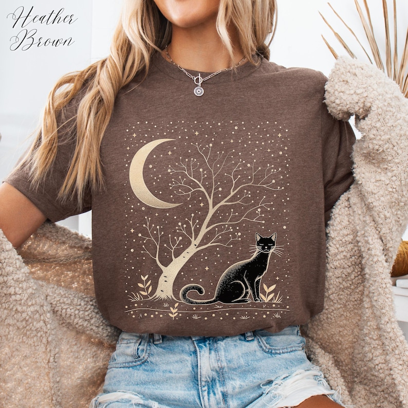 May include: Heather Brown t-shirt featuring a gold crescent moon, tree, and a black cat design. The graphic includes a starry night sky. The shirt is a neutral brown color.