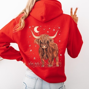 Op de afbeelding: Rode hoodie met een illustratie van een Schotse Hooglandkoe. Het ontwerp toont een bruine koe met lange hoorns, een maan en sterren. De hoodie is gemaakt van een zacht materiaal en is geschikt voor casual wear.