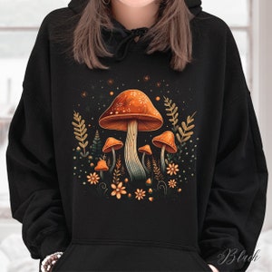Op de afbeelding: Zwarte hoodie met een kleurrijk paddenstoelontwerp. Het ontwerp bevat een grote oranje paddenstoel met witte stippen, kleinere paddenstoelen, bloemen en bladeren. Het woord "Black" staat rechtsonder gedrukt.