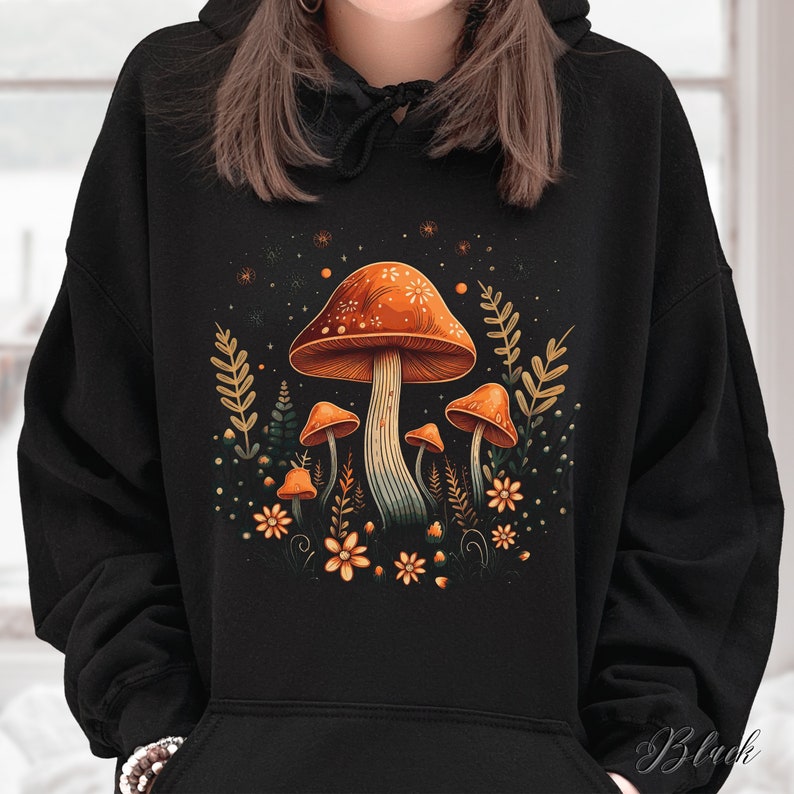 Op de afbeelding: Zwarte hoodie met een kleurrijk paddenstoelontwerp. Het ontwerp bevat een grote oranje paddenstoel met witte stippen, kleinere paddenstoelen, bloemen en bladeren. Het woord "Black" staat rechtsonder gedrukt.