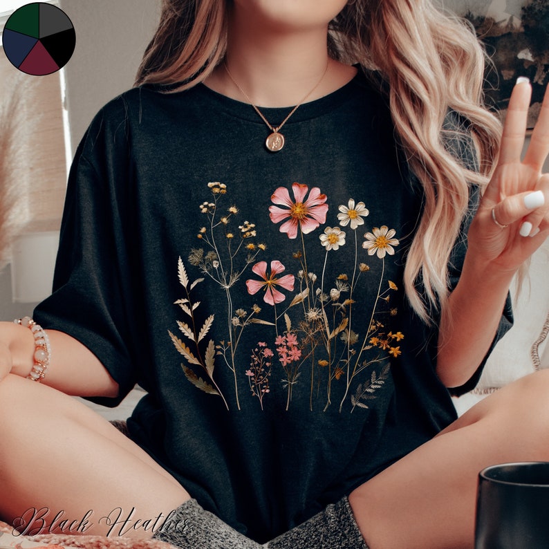 May include: A black t-shirt with a floral print featuring pink, white, and yellow flowers. The flowers are arranged in a cluster and are surrounded by green leaves.