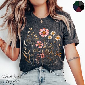 May include: A dark grey heather t-shirt with a floral print of pressed flowers in shades of pink, yellow, and brown. The text "Mon Oc&eacute;an" is written on the wearer's arm.