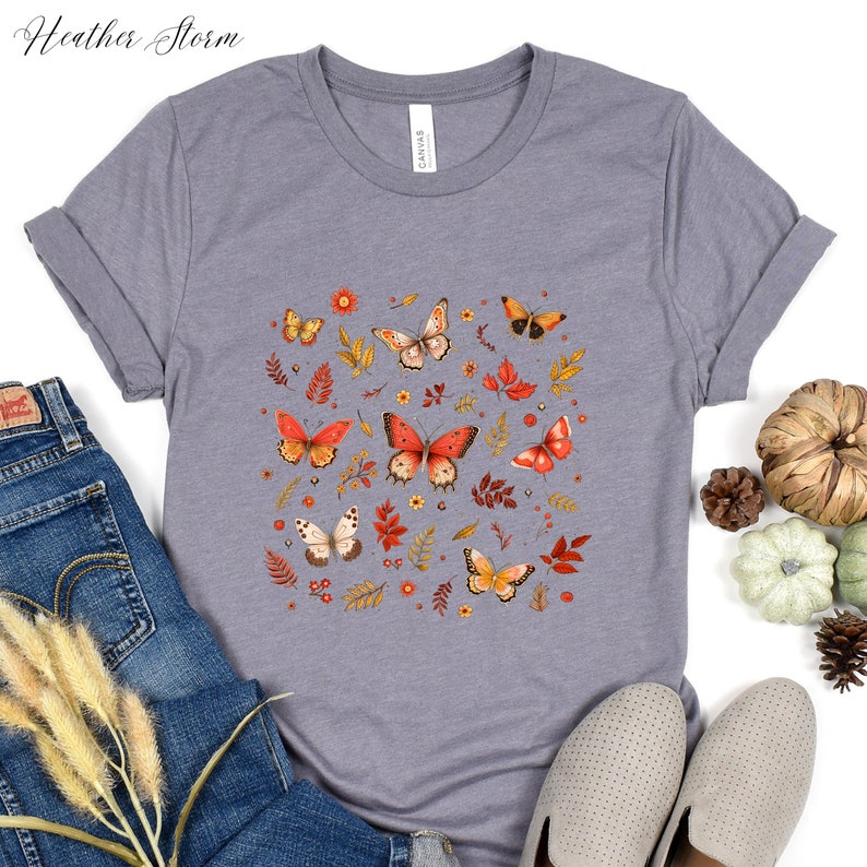 May include: A heather grey t-shirt with a print of orange and yellow butterflies, leaves, and flowers. The design is centered on the shirt.