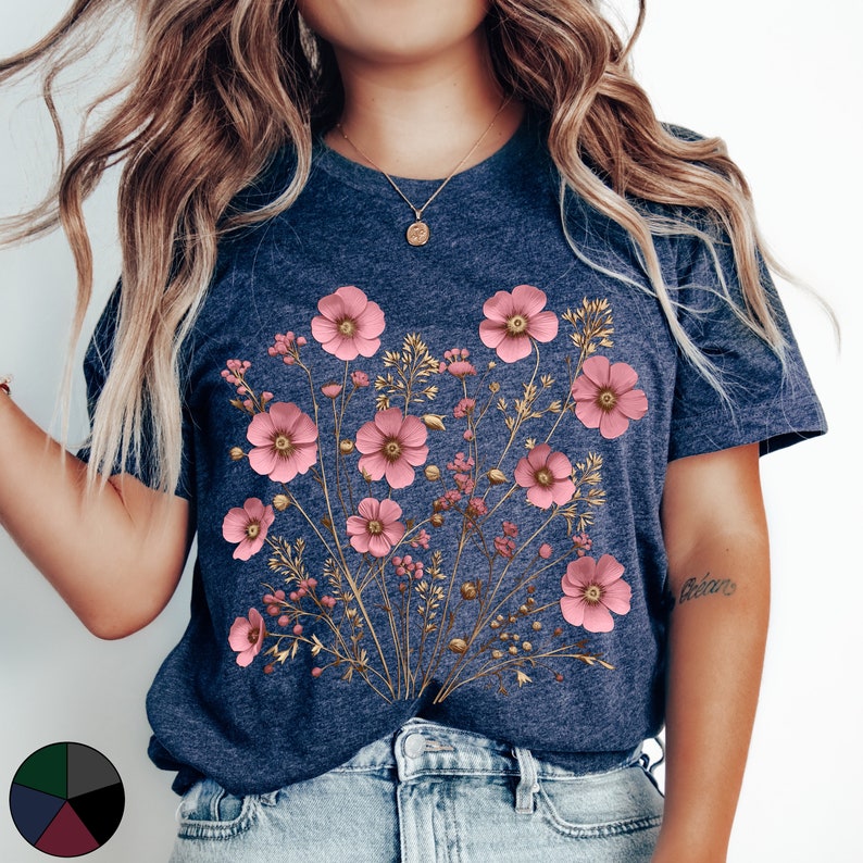 Pu&ograve; includere: Una t-shirt blu navy con una stampa floreale con fiori rosa e steli dorati. I fiori sono disposti a forma di bouquet e sono centrati sulla t-shirt.