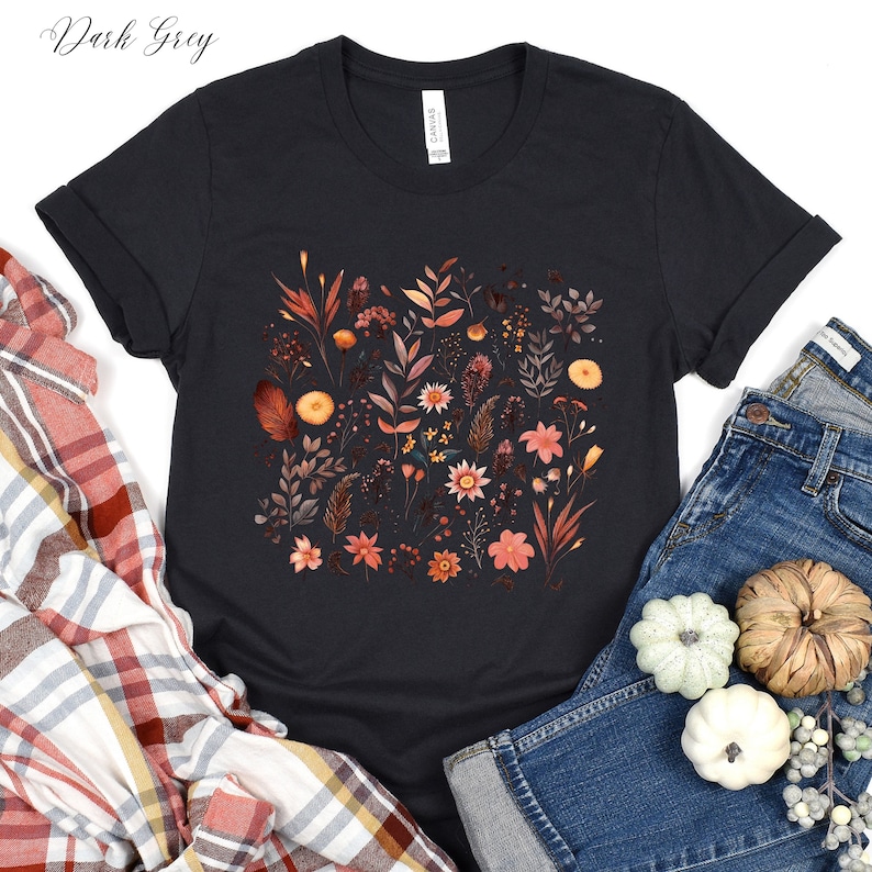 K&ouml;nnte beinhalten: Ein dunkelgraues T-Shirt mit einem floralen Druck, der verschiedene Bl&auml;tter, Bl&uuml;ten und Stiele in Orange-, Braun- und Rosat&ouml;nen zeigt.