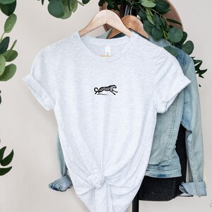 May include: A gray t-shirt with a black and white illustration of a cheetah running. The t-shirt is knotted at the bottom and hanging on a wooden hanger.