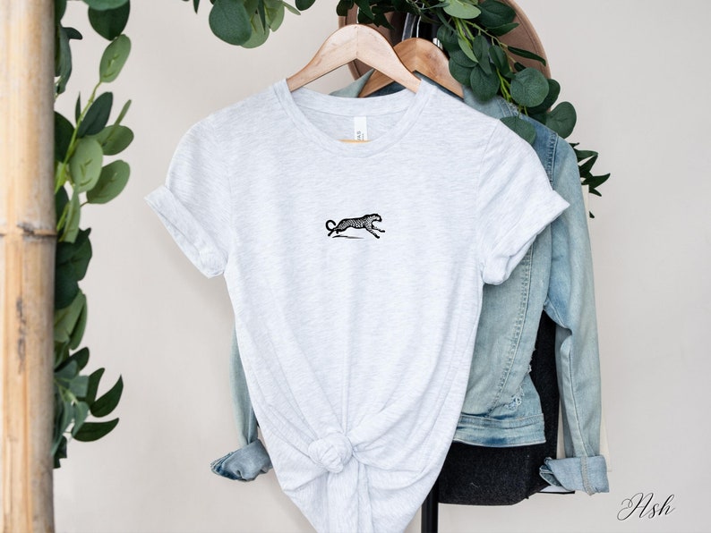 May include: A gray t-shirt with a black and white illustration of a cheetah running. The t-shirt is knotted at the bottom and hanging on a wooden hanger.