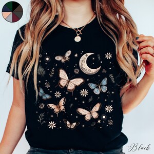 May include: A black t-shirt with a graphic print of butterflies, stars, and a crescent moon. The design is in shades of gold, blue, and white.