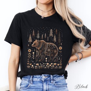May include: Black t-shirt featuring a brown bear with intricate gold-colored patterns. The bear is set against a backdrop of stylized trees and floral elements. The word "Black" is printed at the bottom right.