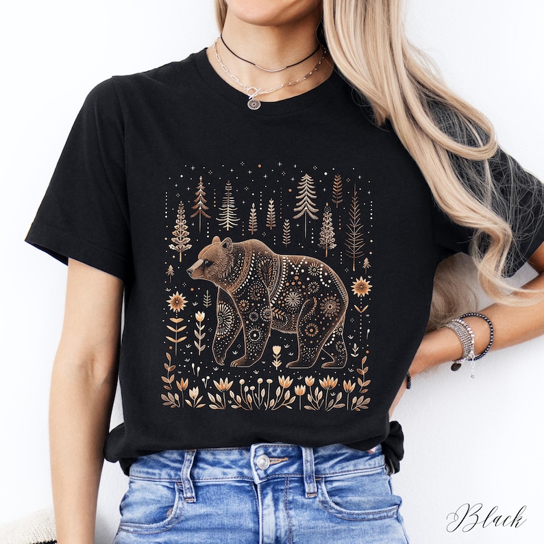 May include: Black t-shirt featuring a brown bear with intricate gold-colored patterns. The bear is set against a backdrop of stylized trees and floral elements. The word "Black" is printed at the bottom right.