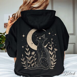 May include: Black hoodie with a graphic print of a black cat sitting under a crescent moon with stars and flowers. The text "Black" is visible in the bottom right corner of the image.