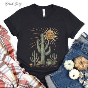 May include: Dark grey t-shirt featuring a desert-themed graphic. The design includes a smiling sun, cacti, and stars. The shirt is paired with blue jeans and decorative gourds.