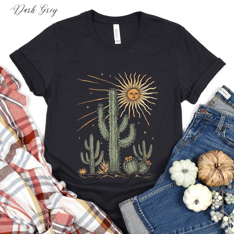 May include: Dark grey t-shirt featuring a desert-themed graphic. The design includes a smiling sun, cacti, and stars. The shirt is paired with blue jeans and decorative gourds.