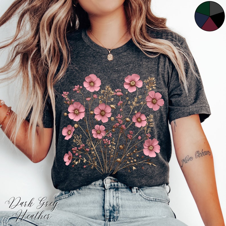 Pu&ograve; includere: Una t-shirt color grigio scuro heather con una stampa floreale con fiori rosa e steli dorati. La t-shirt ha un collo rotondo e maniche corte.