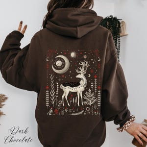 Puede incluir: Una sudadera con capucha marrón con un gráfico de ciervo blanco y luna en la espalda. El gráfico presenta un ciervo estilizado con cuernos, una luna creciente y estrellas en un entorno de bosque. El texto "Dark Chocolate" es visible en la parte inferior de la imagen.