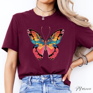 Pu&ograve; includere: T-shirt color bordeaux con un disegno di farfalla colorata. La farfalla presenta tonalit&agrave; rosa, arancioni, gialle e blu. La parola "Maroon" &egrave; stampata in corsivo in basso a destra.