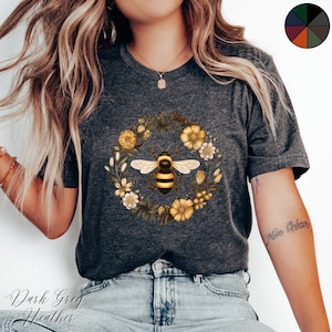 May include: Dark grey heather t-shirt with a bee and floral wreath design in yellow, cream, and brown. The text "Dark Grey Heather" is printed at the bottom of the image. The t-shirt is a crew neck style.