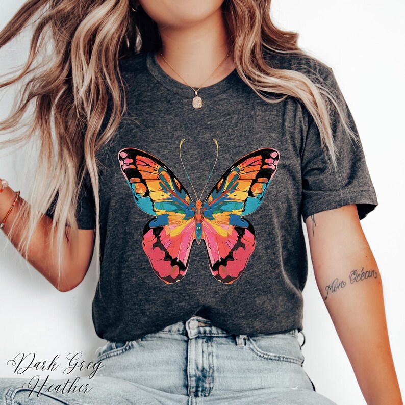 Pu&ograve; includere: T-shirt grigio scuro m&eacute;lange con una grafica a farfalla colorata. Il design della farfalla include sfumature di rosa, arancione, giallo e blu. Il testo "Dark Greg Heather" &egrave; stampato in basso sulla maglietta.