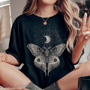 May include: Black t-shirt with a graphic of a moth with a crescent moon above it. The moth has large, detailed wings and a textured body.