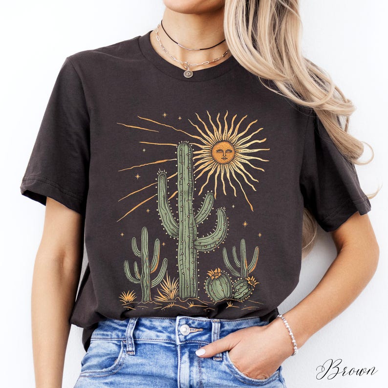 May include: A dark brown t-shirt featuring a desert-themed graphic. The design includes a sun with a face, cacti, and stars. The word "Brown" is written in cursive in the bottom right corner.