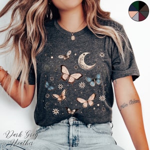 May include: A dark grey heather t-shirt with a crescent moon and butterflies printed on the front. The butterflies are in shades of blue, pink, and gold. The text "Mon Oc&eacute;an" is written on the wearer's arm.
