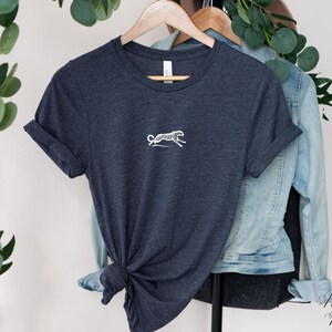 May include: A heather navy blue t-shirt with a white embroidered cheetah design. The t-shirt is folded and displayed on a hanger.