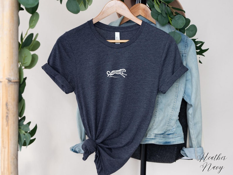 May include: A heather navy blue t-shirt with a white embroidered cheetah design. The t-shirt is folded and displayed on a hanger.