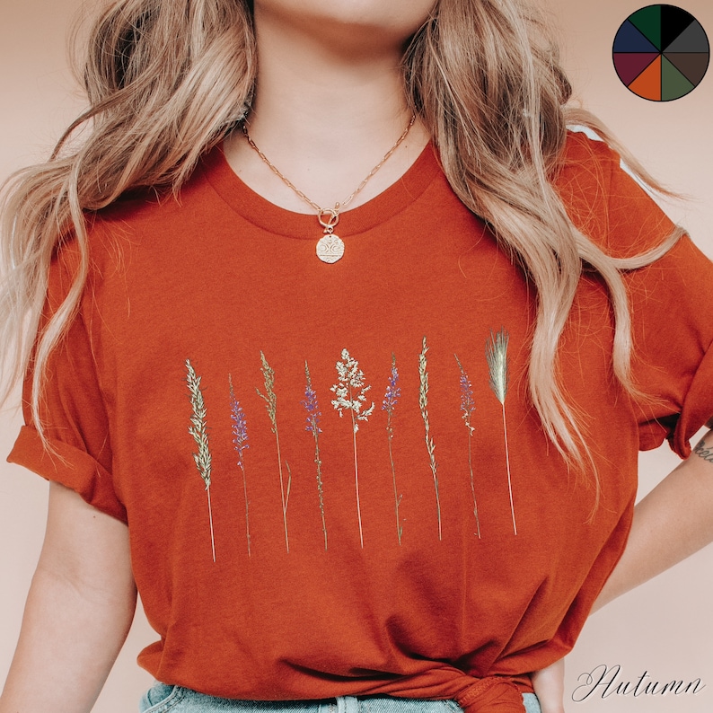 May include: A rust-coloured t-shirt with a graphic of dried flowers and grasses. The shirt has a relaxed fit and short sleeves.