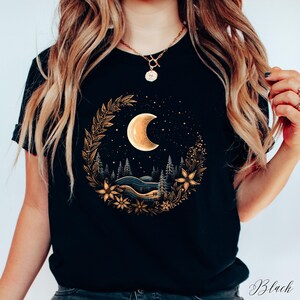 May include: Black t-shirt with a gold crescent moon and floral design. The moon is surrounded by a wreath of leaves and flowers. The design is centered on the shirt.