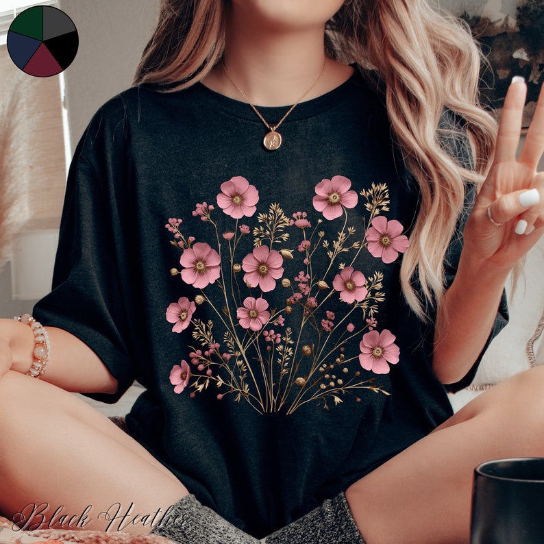 Pu&ograve; includere: Una maglietta nera con una stampa floreale con fiori rosa e steli dorati. I fiori sono disposti a forma di bouquet.