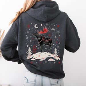 May include: Dark gray hoodie featuring a winter scene with a black moose with red antlers and a decorative blanket. The design includes snowflakes, a crescent moon, and two pine trees. The moose stands on a snowy hill.