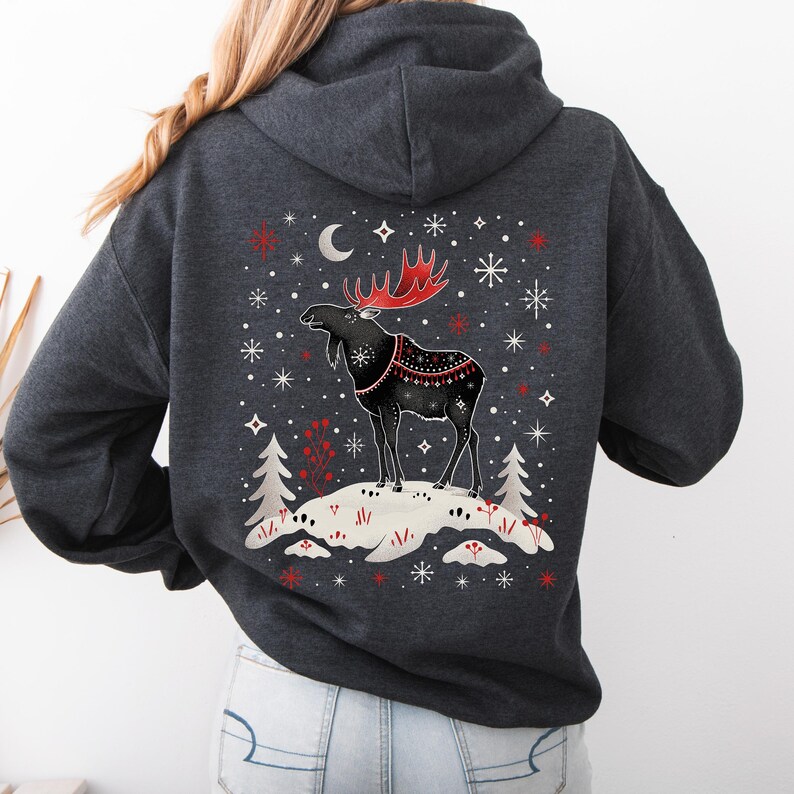 May include: Dark gray hoodie featuring a winter scene with a black moose with red antlers and a decorative blanket. The design includes snowflakes, a crescent moon, and two pine trees. The moose stands on a snowy hill.