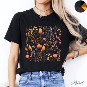 Może przedstawiać: Czarny T-shirt z nadrukiem kwiatowym przedstawiającym r&oacute;żne kwiaty i liście w odcieniach pomarańczowego, ż&oacute;łtego i r&oacute;żowego. Nadruk jest umieszczony centralnie na koszulce i pokrywa większość przodu.