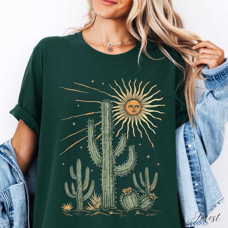 May include: Dark green t-shirt featuring a desert-themed graphic. The design includes a sun with a face, cacti, and stars. The shirt has rolled-up sleeves and a relaxed fit.