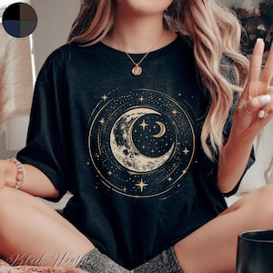 Puede incluir: Una camiseta negra con un gráfico de luna creciente y estrellas doradas. La luna tiene una superficie texturizada y está rodeada por un círculo de estrellas.