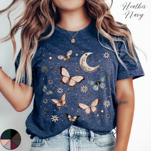 May include: A heather navy blue t-shirt with a graphic print of butterflies, a crescent moon, and stars. The shirt has a crew neck and short sleeves.