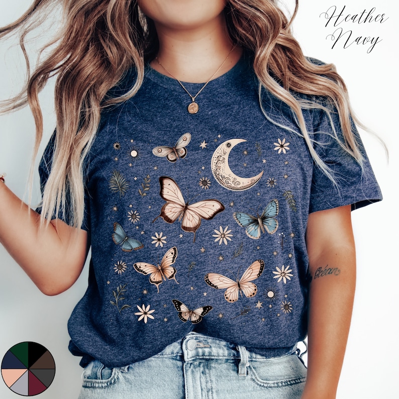 May include: A heather navy blue t-shirt with a graphic print of butterflies, a crescent moon, and stars. The shirt has a crew neck and short sleeves.