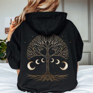 Puede incluir: Sudadera con capucha negra con un diseño de árbol de la vida dorado en la espalda. El árbol tiene ramas y raíces intrincadas y está rodeado de lunas crecientes.