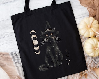 May include: Black tote bag featuring a black cat wearing a witch hat, with a crescent moon design and stars. The cat has yellow eyes and white whiskers. The bag has black handles and is likely made of canvas or a similar material.
